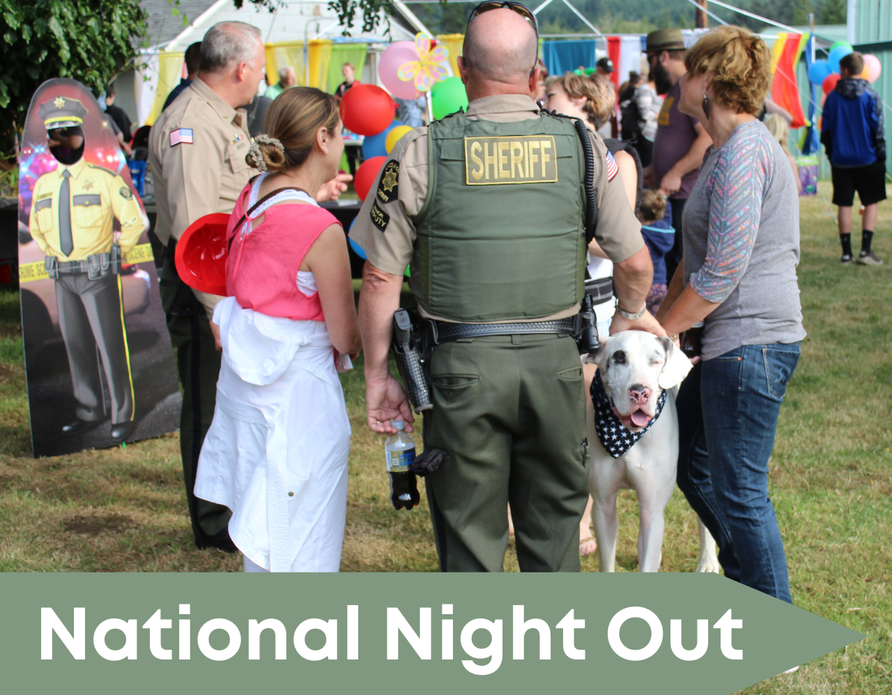 People at a community event standing with their backs to camera. Deputy in uniform petting a dog. 
